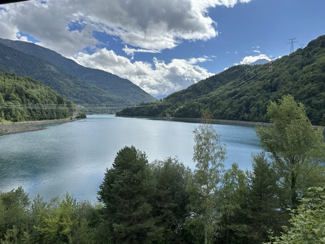 Near Allemond, France. View on Lac du Verney. A row of trees in the foreground, an expansive lake in the middle, trees and mountains to the left and right of the lake, a stream appears above the lake in the distance.
